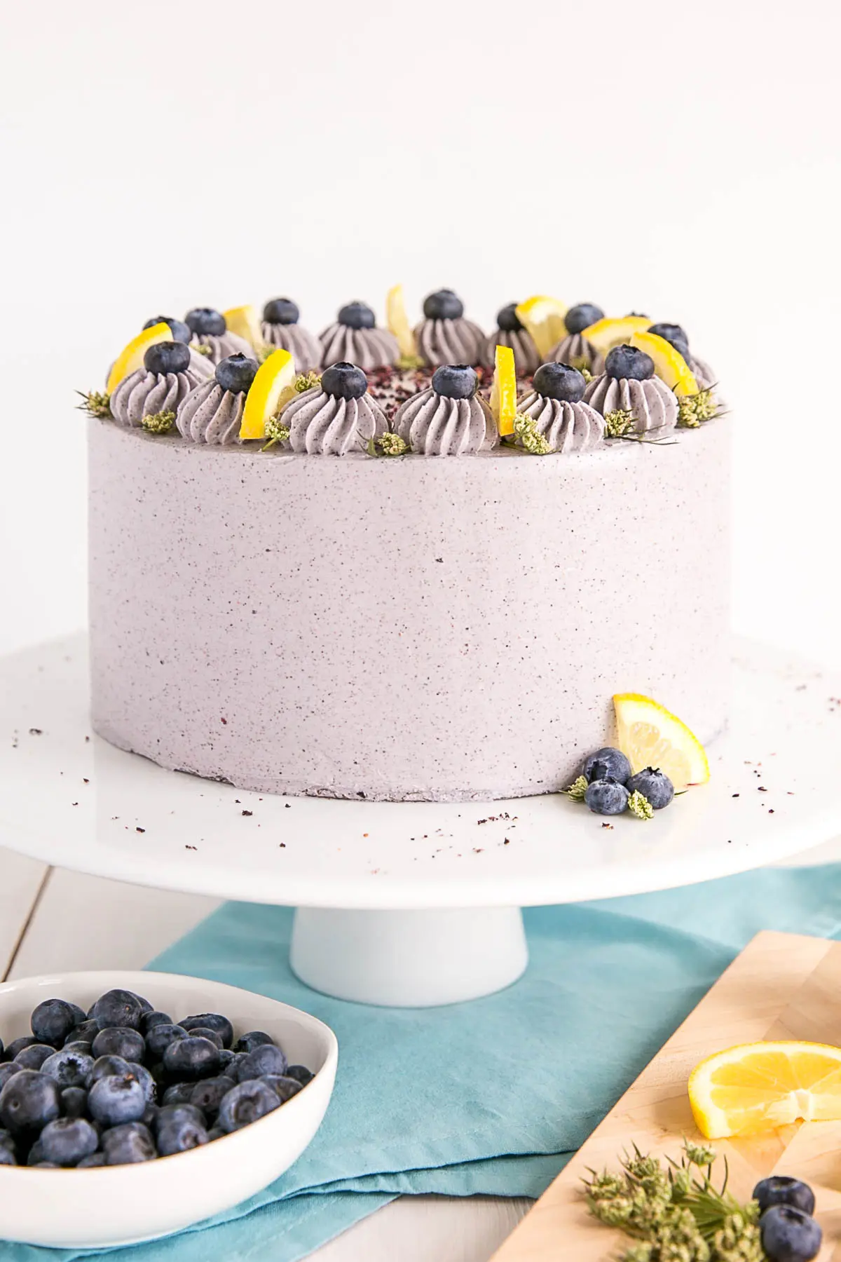 Lemon Blueberry Cake on a white cake stand decorated with lemon wedges and blueberries.