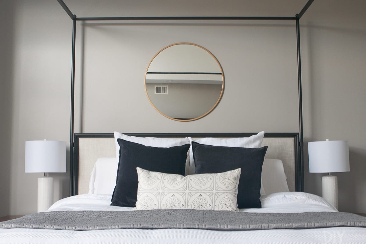 Queen size master bedroom bed with white sheets, black and white pillows, and a round gold detailed mirror hanging over the bed. 