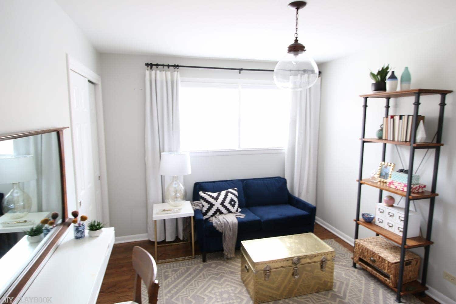 A bright, neutral colored sitting area in the master bedroom with a bookshelf, blue loveseat, and sleek vanity area. 