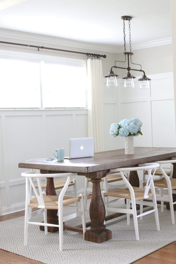 Rustic dining room table with white wooden chairs and hanging light fixture. 