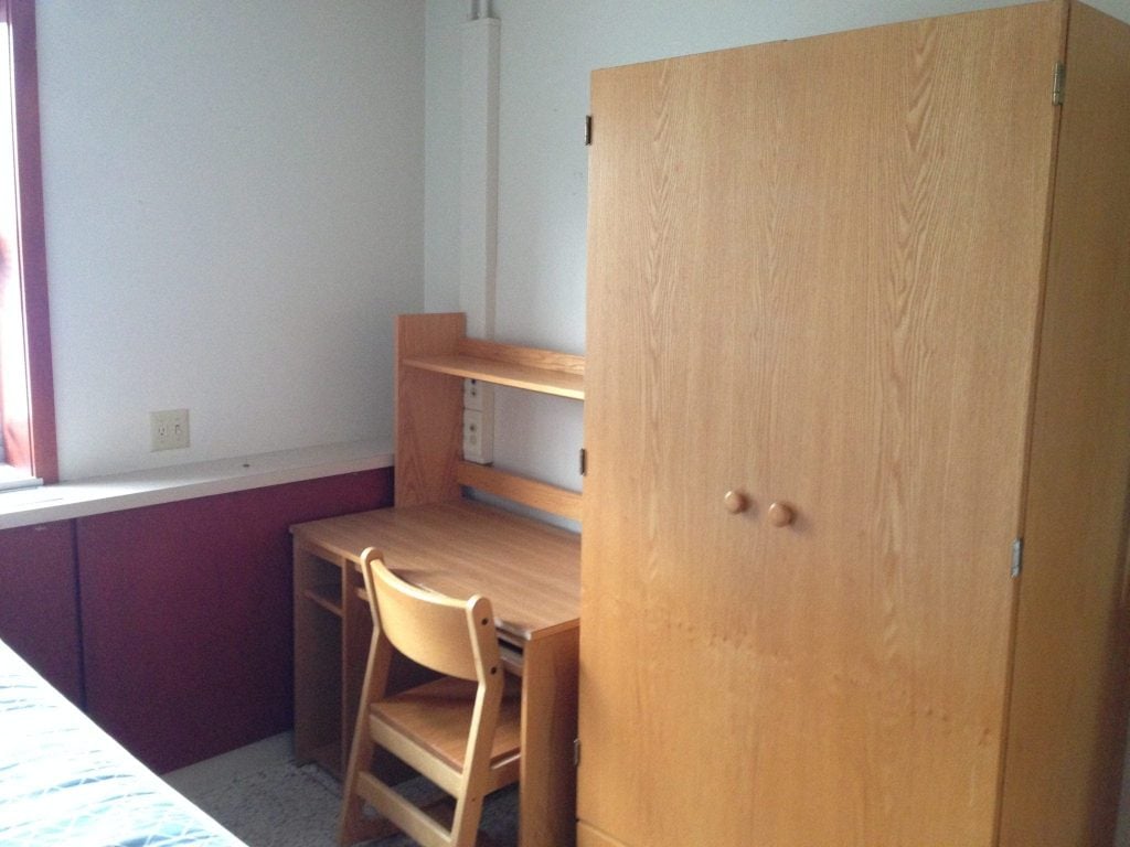 A tiny dorm room with a bed, desk and large dresser. 